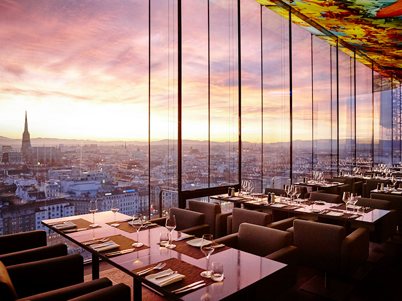 Empty tables in a rooftop restaurant beside a large window at sunset over the city