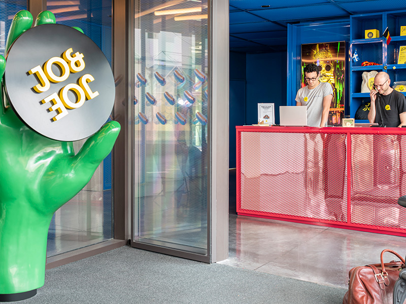 Two people stand on phones behind a red check-in desk, with a giant green hand holding the Jo&Joe logo on the left