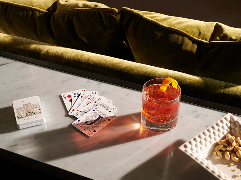 A negroni sits on a table beside a pack of playing cards and plate of peanuts