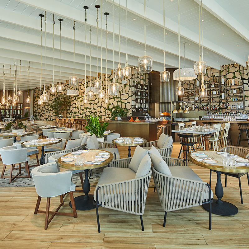 A restaurant set up for tables of 4-6 guests, It is well lit by hanging lights coming down from the ceiling. There is a bar in the background and a striking wall design behind that.